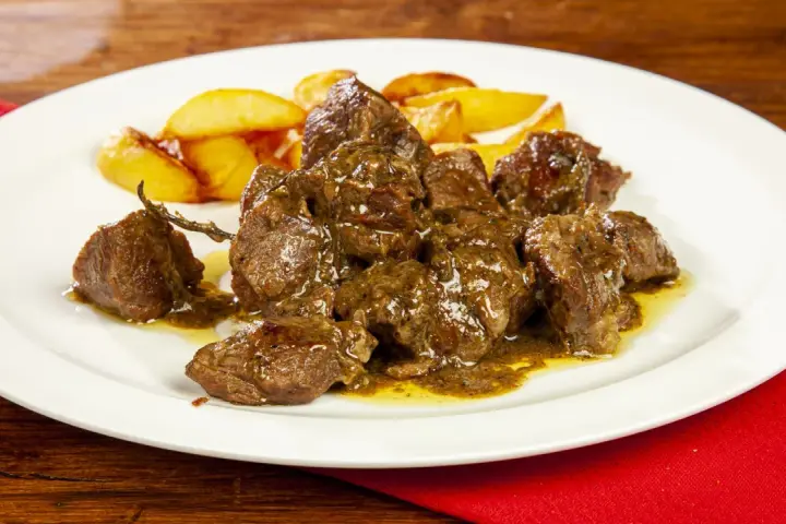 Un saporito abbacchio alla cacciatora, tenero e succulento, servito con patate arrosto su un piatto bianco.