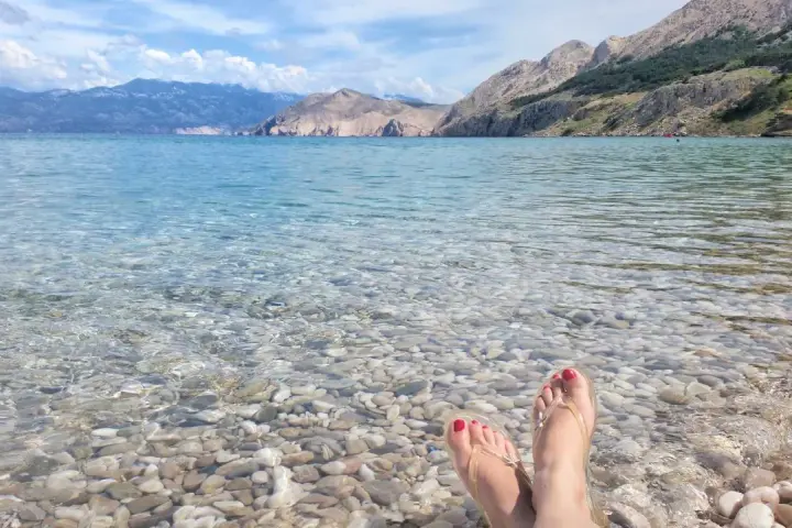 Stopy w klapkach na kamienistej plaży w **Krk National Park**. Woda krystalicznie czysta, w tle góry.