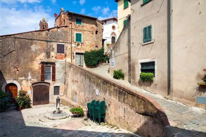 Un vicolo pittoresco a Castagneto Carducci, con case antiche, un fontanello e sedie verdi impilate.