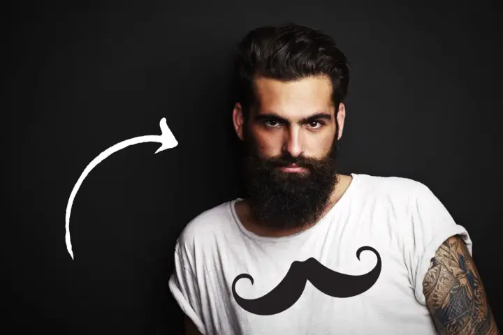 Hombre con barba tupida, quizás decidiendo dejarse barba por primera vez. Camiseta blanca con estampado de bigote.