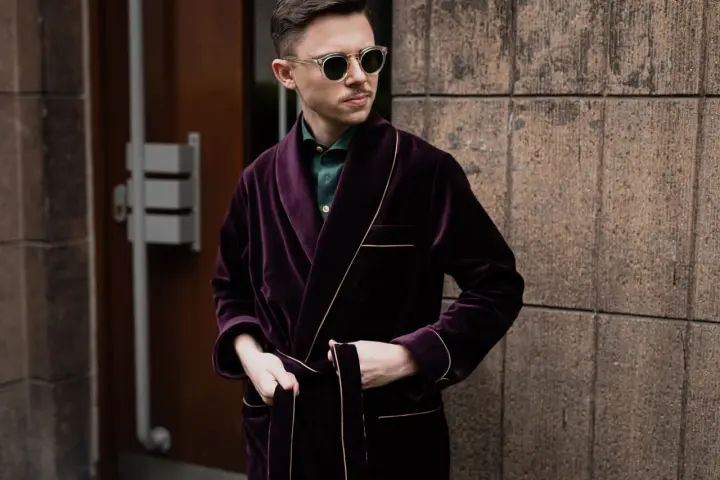 Elegancki mężczyzna w bordowej bonżurce z aksamitu, w okularach przeciwsłonecznych, stoi przy kamiennej ścianie.
