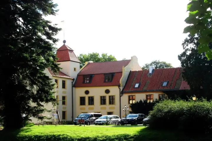 Zamek z czerwoną wieżą i dachami, otoczony zielenią. Samochody zaparkowane na trawniku. Idealne miejsce na krokowa atrakcje.