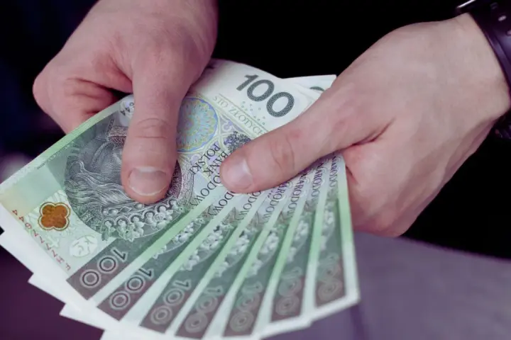 Dłonie trzymają stos banknotów 100 zł. Czy to wystarczy, by pokryć ile wynosi najniższa krajowa?