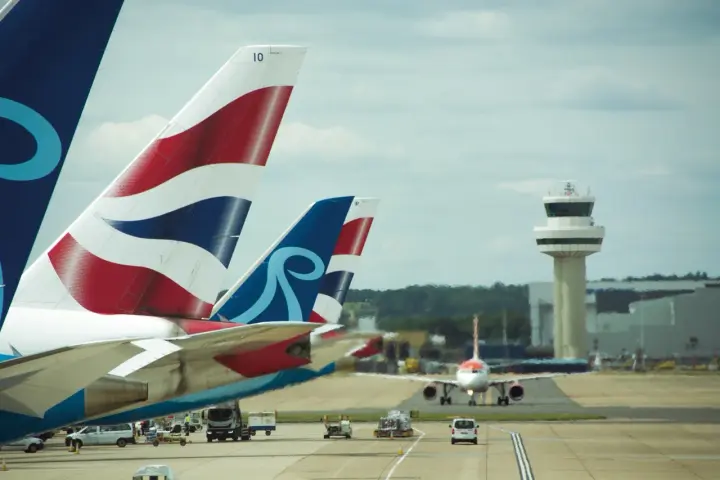 Samoloty z charakterystycznymi ogonami British Airways i innym logo czekają na start na lotnisku Londyn Gatwick. W tle wieża kontroli lotów.