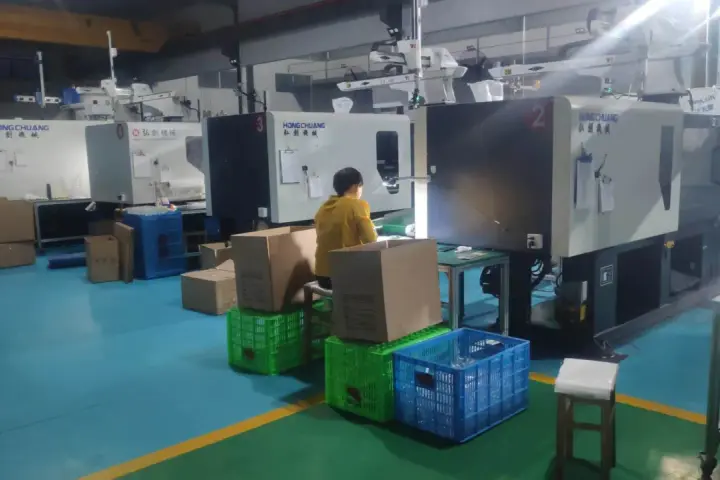 A person in a yellow jacket works at a station with boxes and crates, surrounded by injection molding machines, indicating low volume production.