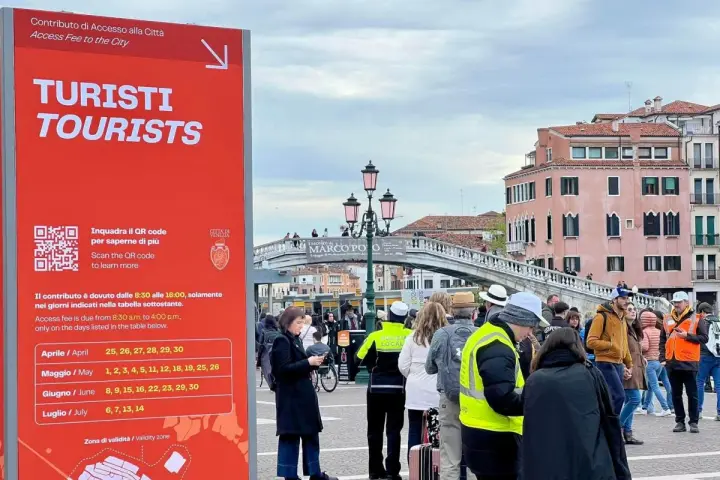 Cartello rosso a Venezia per il contributo accesso turisti, con date e QR code. Persone in fila sullo sfondo.