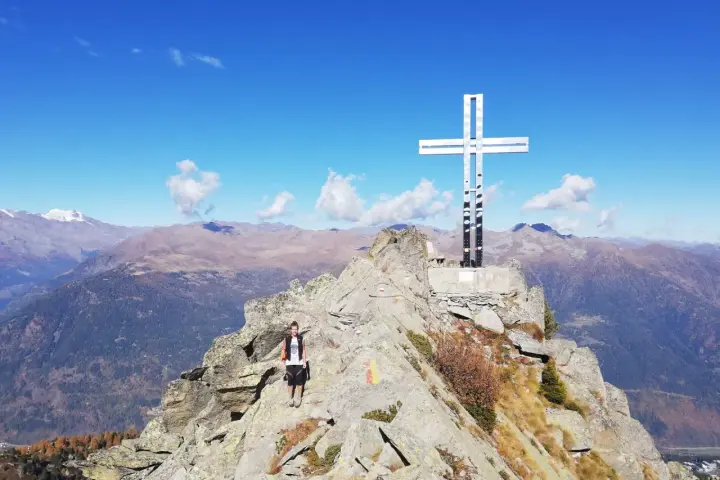 Escursionista raggiunge la vetta con la croce della pace, circondata da montagne maestose sotto un cielo azzurro.