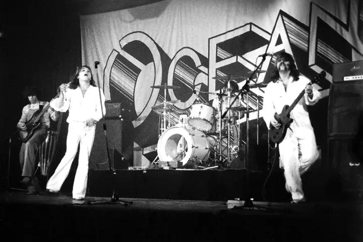 Le groupe Trust en concert, avec le chanteur en blanc au centre, un guitariste à gauche et un autre guitariste à droite. La scène est décorée d'une grande bannière avec le logo du groupe.