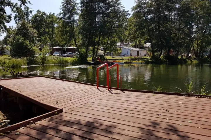 Drewniany pomost z czerwonymi poręczami nad jeziorem. W tle drzewa, budynki i samochody. Idealne miejsce na łagów camping.