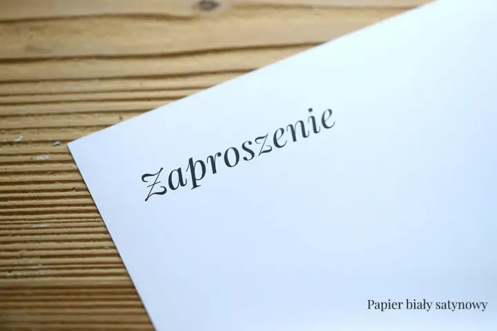 Eleganckie zaproszenie na ślub na białym, satynowym papierze, leżące na drewnianym tle.