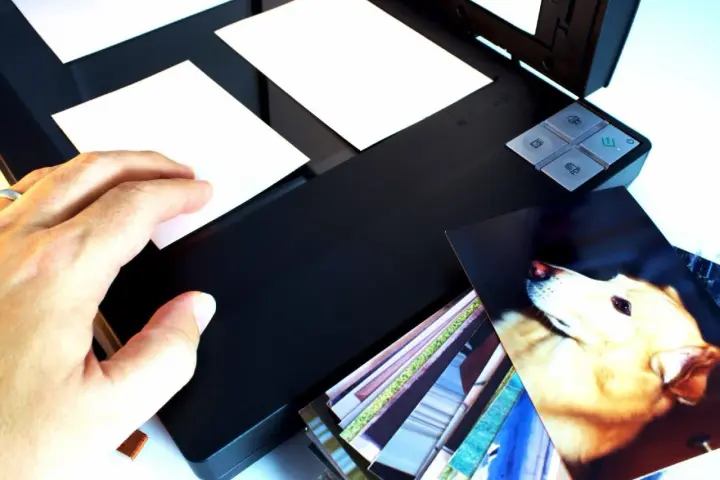 Ręka kładzie zdjęcie na skanerze, rozpoczynając proces digitalizacji zdjęć. Obok stos innych fotografii.