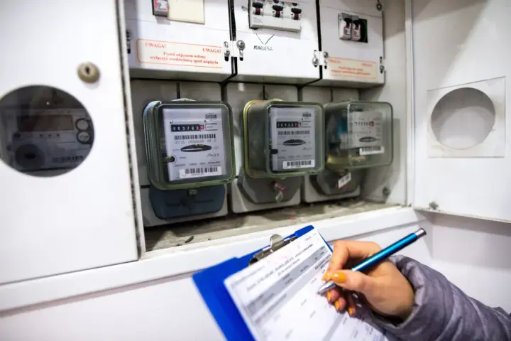 Kontrola liczników energii elektrycznej. Ręka z długopisem zapisuje dane na kartce, obok widoczne liczniki i bezpieczniki.