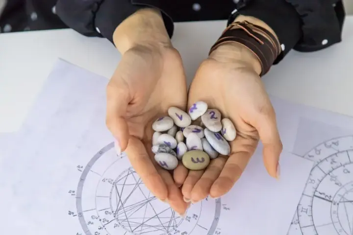 Dłonie trzymają kamyki z numerami, jeden z nich to 3, co może symbolizować numerologiczną 6. W tle wykresy astrologiczne.