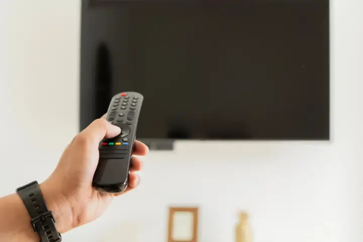 Dłoń trzymająca pilot do telewizora na tle wyłączonego ekranu.