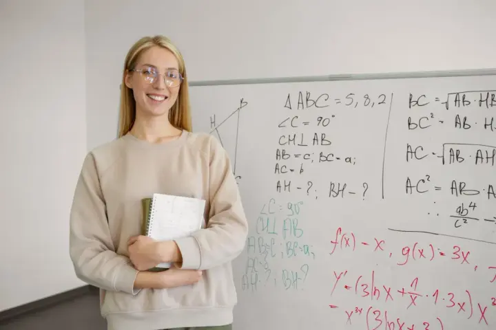 Uśmiechnięta nauczycielka z zeszytem przy tablicy z matematycznymi wzorami. Zastanawia się, ile punktów na maturze z matematyki można zdobyć za takie zadania.
