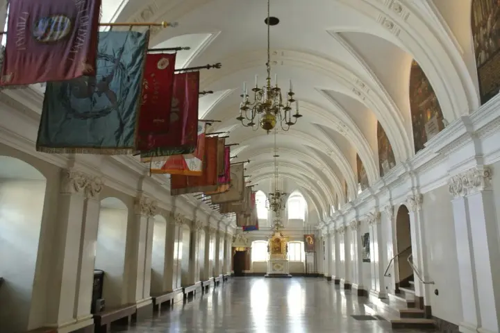 Długi korytarz z łukami i żyrandolami, ozdobiony historycznymi sztandarami. To wnętrze klasztoru na Jasnej Górze, gdzie każdy baner opowiada historię zakonu paulinów.