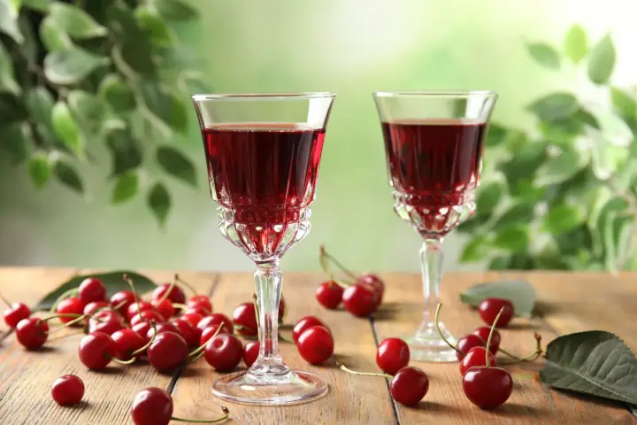Dwa kieliszki z rubinową nalewką z wiśni na spirytusie, obok świeże wiśnie na drewnianym stole. Szybka nalewka z wiśni na spirytusie gotowa do degustacji.