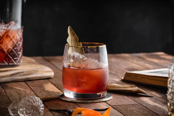 Czerwony koktajl z lodem i liściem laurowym w szkle, obok karafki i starej książki. To może być drink z wermutem.