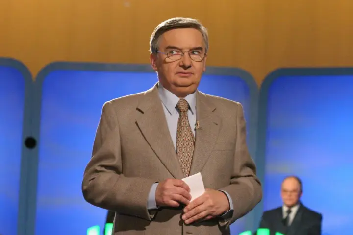 Tadeusz Sznuk, który prowadzi teleturniej Jeden z dziesięciu, w beżowej marynarce i z kartką w dłoni.