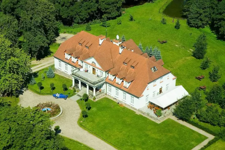 Dwór w Bychowie otoczony zielenią, z niebieskim kabrioletem przy różanym klombie.