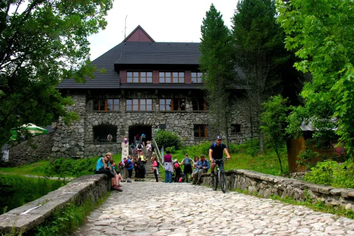 Kamienny budynek schroniska w Dolinie Chochołowskiej z ludźmi na schodach i rowerzystą na kamiennej drodze.