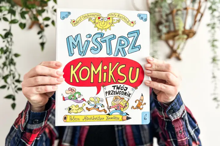 Okładka książki "Mistrz Komiksu" z rysunkami postaci i słońca, trzymana przez ręce.