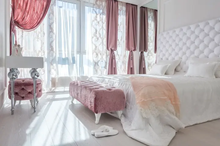 Elegancka sypialnia w stylu angielskim z pikowanym zagłówkiem, różowymi pufami i zasłonami.