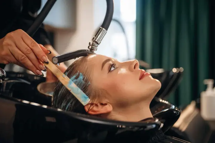 Profesjonalna pielęgnacja włosów farbowanych w salonie. Stylistka nakłada maskę na mokre włosy klientki.