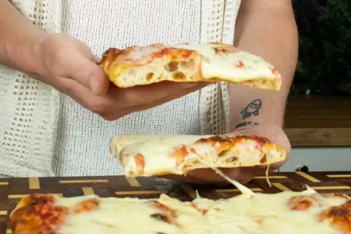 Due fette di pizza in teglia, con mozzarella filante e crosta dorata, vengono sollevate da mani.