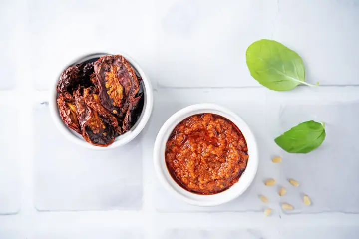 Due ciotoline bianche: una con pomodori secchi interi, l'altra con un invitante pesto di pomodori secchi. Foglie di basilico e pinoli completano la scena.