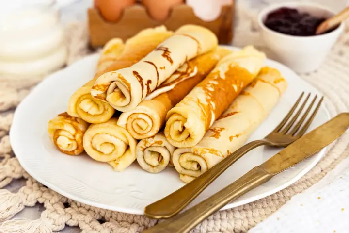 Ein Teller mit goldbraunen Quark Pfannkuchen, aufgerollt und gestapelt. Daneben liegen ein goldenes Messer und eine Gabel. Im Hintergrund eine Schale Marmelade.
