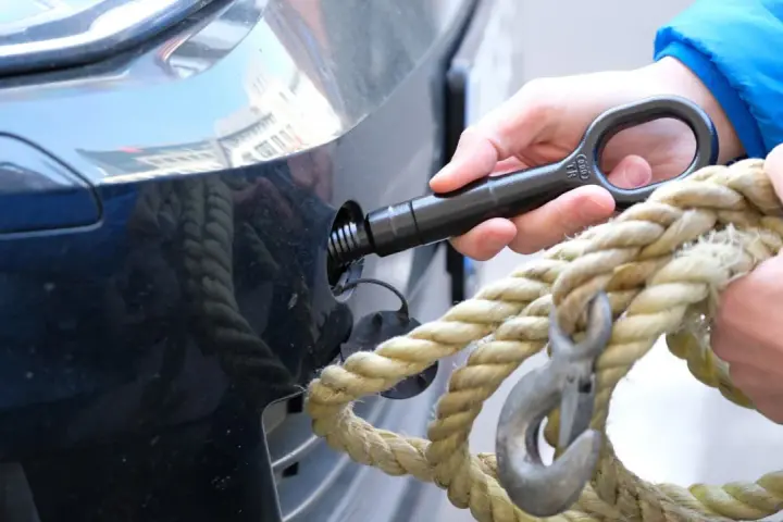 Eine Hand schraubt den Abschlepphaken in das Auto, während eine andere Hand ein dickes Seil mit Haken hält.