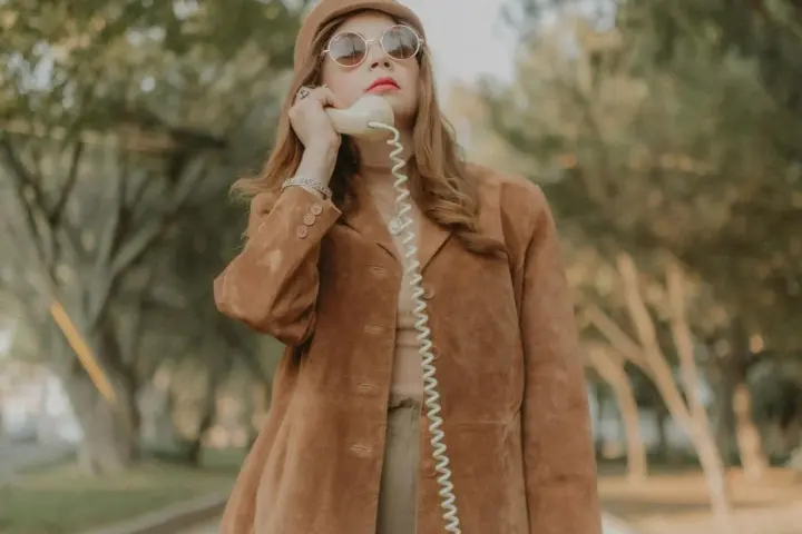Kobieta w brązowej zamszowej kurtce damskiej rozmawia przez telefon w stylu retro.
