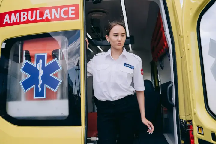 Młoda kobieta w białej koszuli z naszywką "PARAMEDICS" stoi przy otwartych drzwiach karetki. Czy kierowca karetki musi być ratownikiem?
