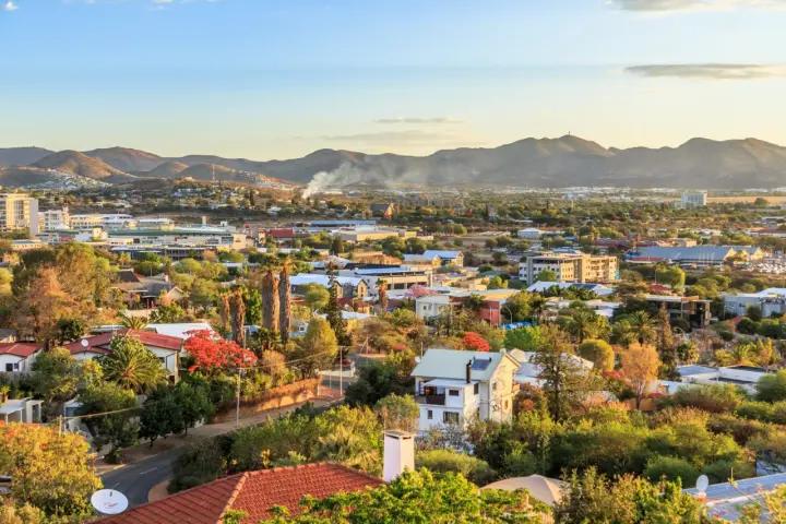 Widok na miasto Windhoek w Namibii o zachodzie słońca. W oddali góry, a bliżej zabudowania mieszkalne i komercyjne, otoczone bujną zielenią.