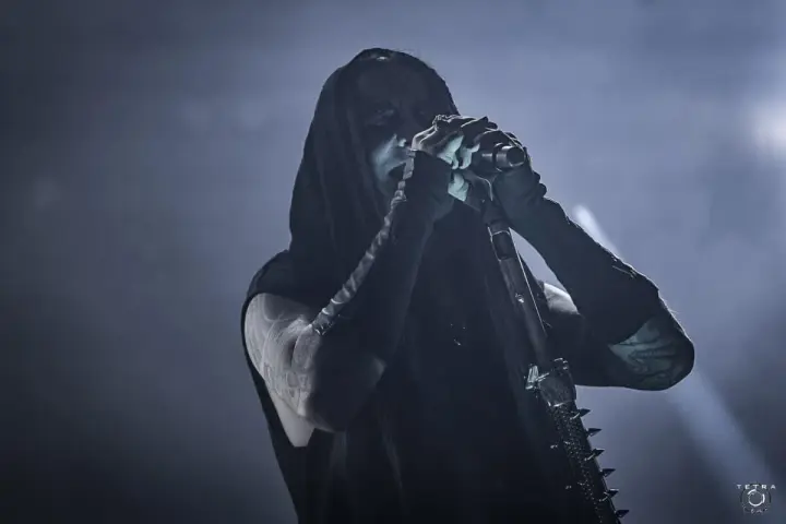 Chanteur de Dimmu Borgir, visage encapuchonné et maquillé, chantant dans un micro, ambiance sombre et enfumée.