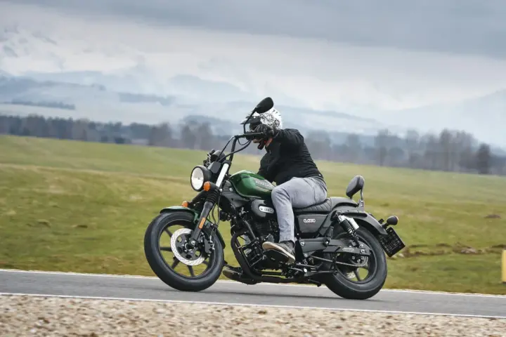 Motocyklista na zielonym chopperze UM Renegade 125 przemierza malowniczą drogę wśród zielonych pól i gór.