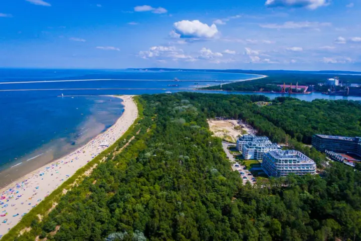 Widok z lotu ptaka na najszerszą plażę w Polsce, pełną wypoczywających ludzi, otoczoną lasem i nowoczesnymi budynkami.