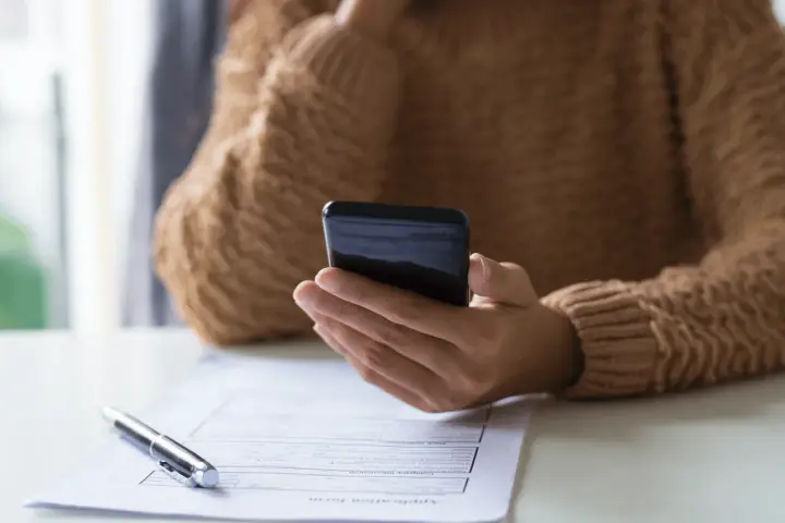Persona revisa su teléfono móvil, modelo 035, mientras tiene un bolígrafo y un formulario de solicitud sobre la mesa.