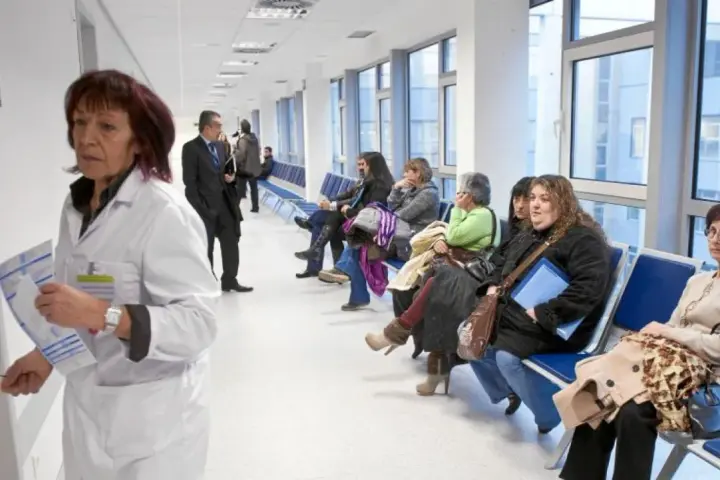 Doctora con bata blanca revisa papeles, mientras pacientes esperan en pasillo. Quizás pensando en cómo pedir excedencia estando de baja.