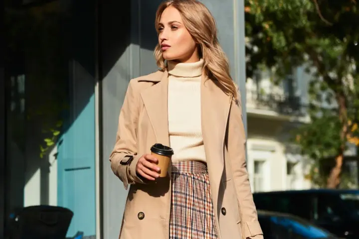 Elegancka kobieta w beżowym płaszczu, kremowym golfie i spódnicy w kratę, z kubkiem kawy w dłoni. Inspiracja na stylizacje do beżowego płaszcza.