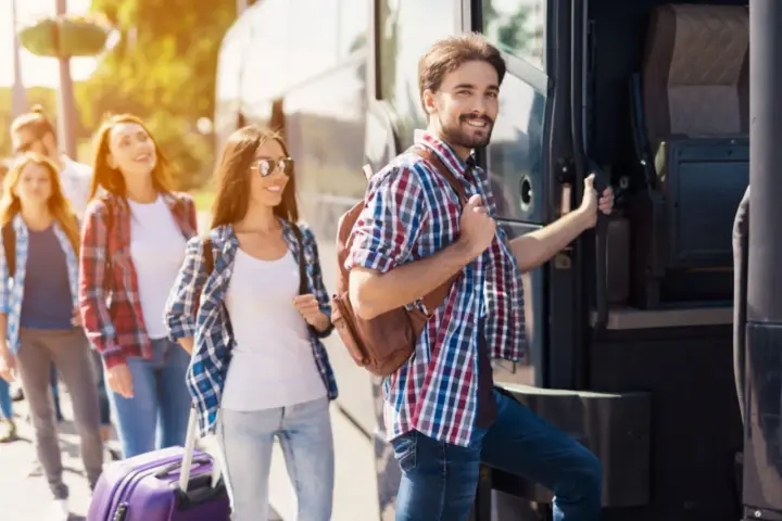 Grupa młodych ludzi z plecakami i walizką wsiada do autobusu, gotowa na wycieczki fakultatywne.