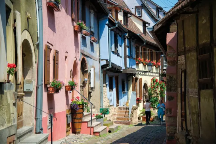 Charming street with colorful houses, perfect for day trips from Strasbourg. Two women stroll past flower-filled window boxes.
