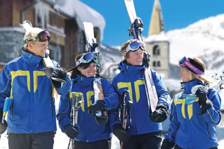 Quatre jeunes skieurs souriants en tenue bleue et jaune, portant des skis et des bâtons, dans une station de ski enneigée.