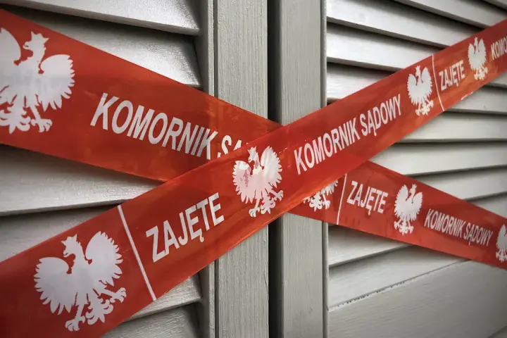 Czerwona taśma z napisem "KOMORNIK SĄDOWY" i godłem Polski krzyżuje się na szarych drzwiach. Nakaz zapłaty z sądu kiedy komornik może oznaczać zajęcie.