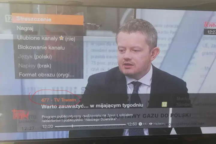 Mężczyzna w garniturze na ekranie telewizora, na którym widać menu z opcją "477 - TV Trwam".
