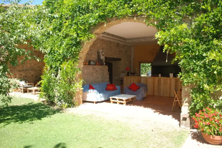 Un jardín con una madreselva planta cubriendo un arco de piedra. Dentro, sofás y una barra de madera.