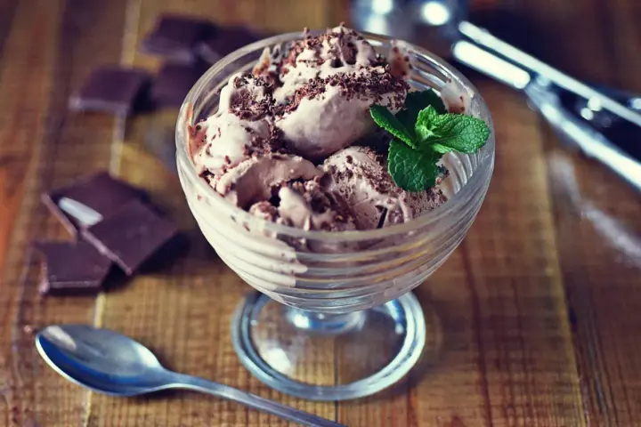 Pyszne lody nuii w pucharku, posypane czekoladą i udekorowane miętą. Obok leżą kawałki czekolady i łyżeczka.