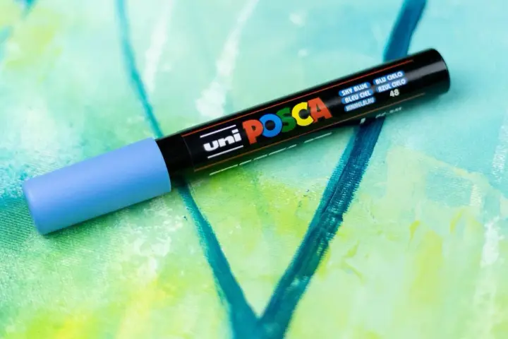 En ljusblå Posca-tuschpenna ligger på en abstrakt målning i gröna och blå nyanser. Perfekt för att måla med tuschpennor.