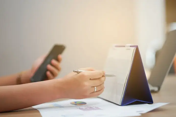 Ręce planują zadania na kalendarzu, trzymając telefon. Zapewnia to ciągłość pracy i organizację dnia.